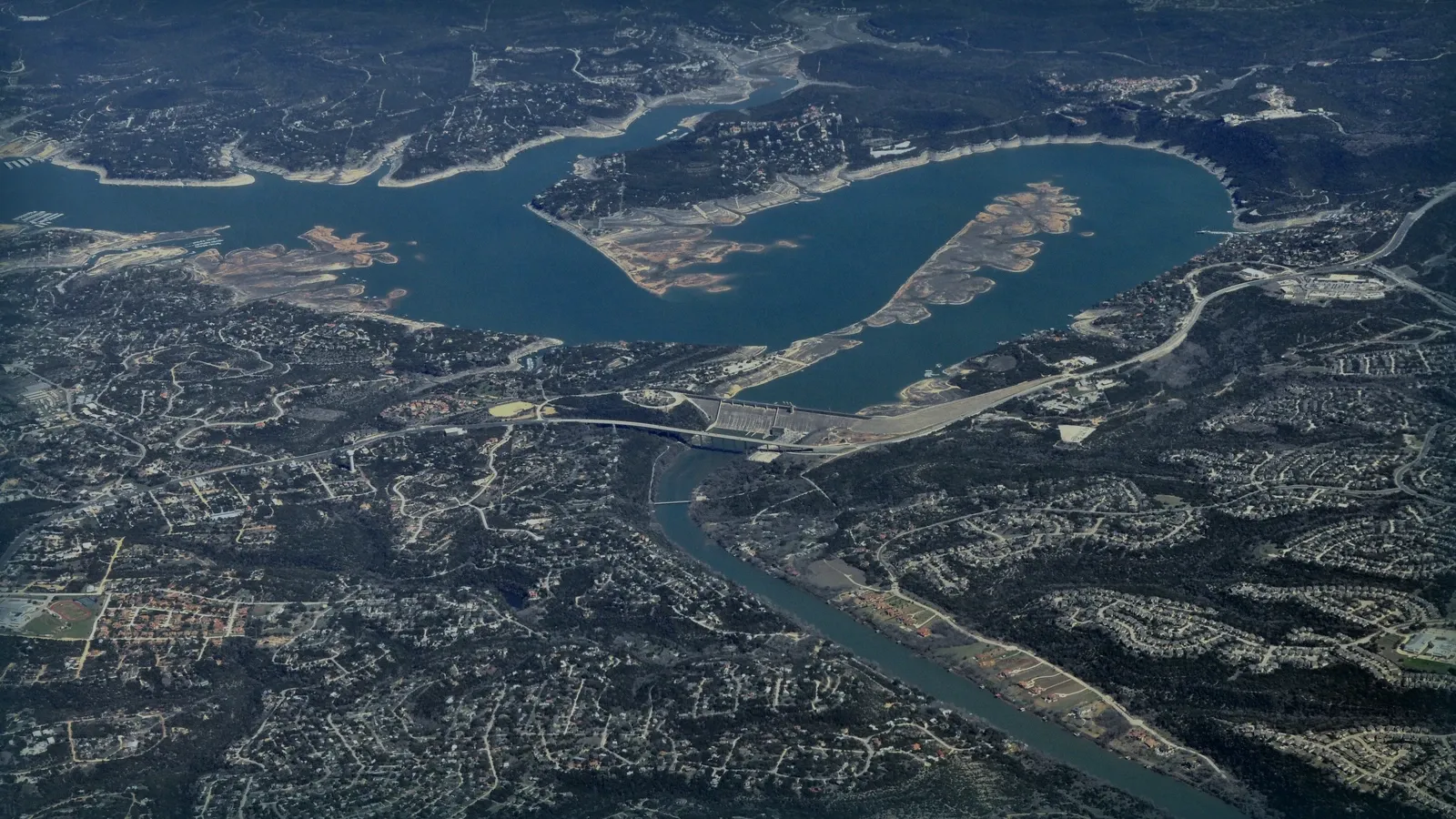 Vista aérea de una presa y su embalse entre laderas urbanizadas.